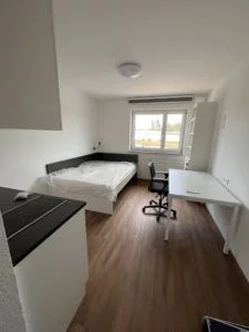 Blick in ein Zimmer, links Pantryküche, oben links Bett, geradeaus Fenster, rechts weißer Schreibtisch mit schwarzem Stuhl