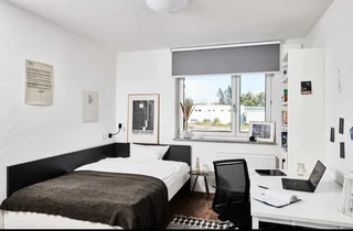 Blick in ein Zimmer, links Bett mit dunkler Tagesdecke, geradeaus Fenster, rechts weißer Schreibtisch mit schwarzem Stuhl