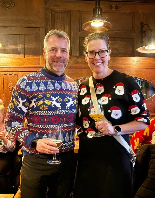 Sebastian Funke und Mnady Bressel mit Weihnachtspullovern und einem Getränk in der Hand